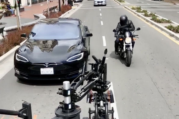 Black Tesla car parked on the side of the road with a motorcycle nearby, street and parking lot in background.