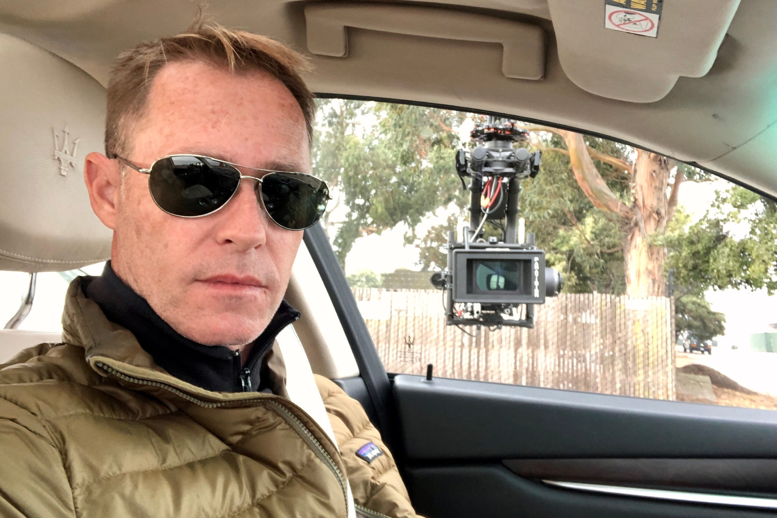 Man wearing sunglasses and a brown jacket sitting in a car, with a camera mounted on the windshield, outdoors.