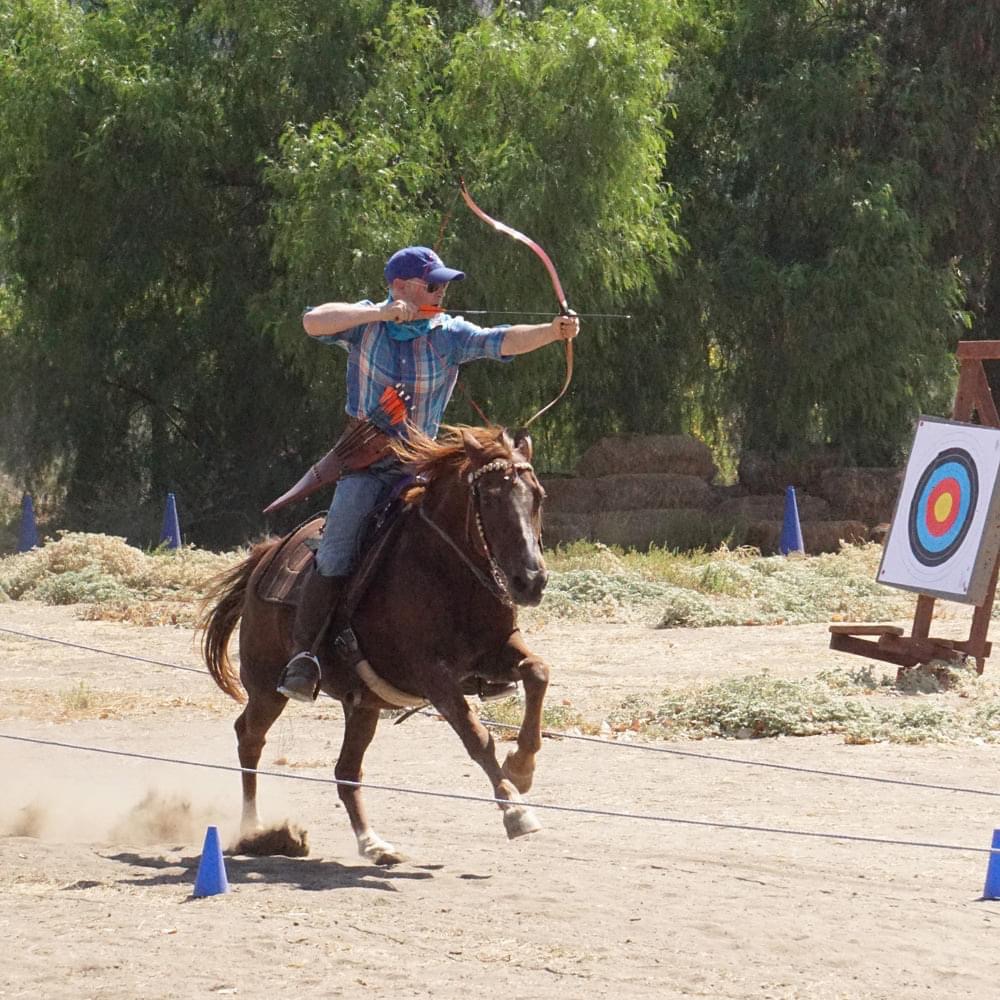Child riding a horse, aiming a bow and arrow outdoors with trees and a target in the background.