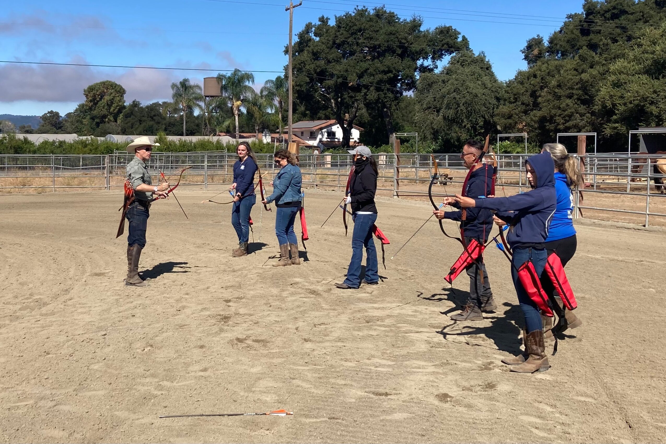 Group of people practicing lassoing on a dirt field with trees and a fence in the background.