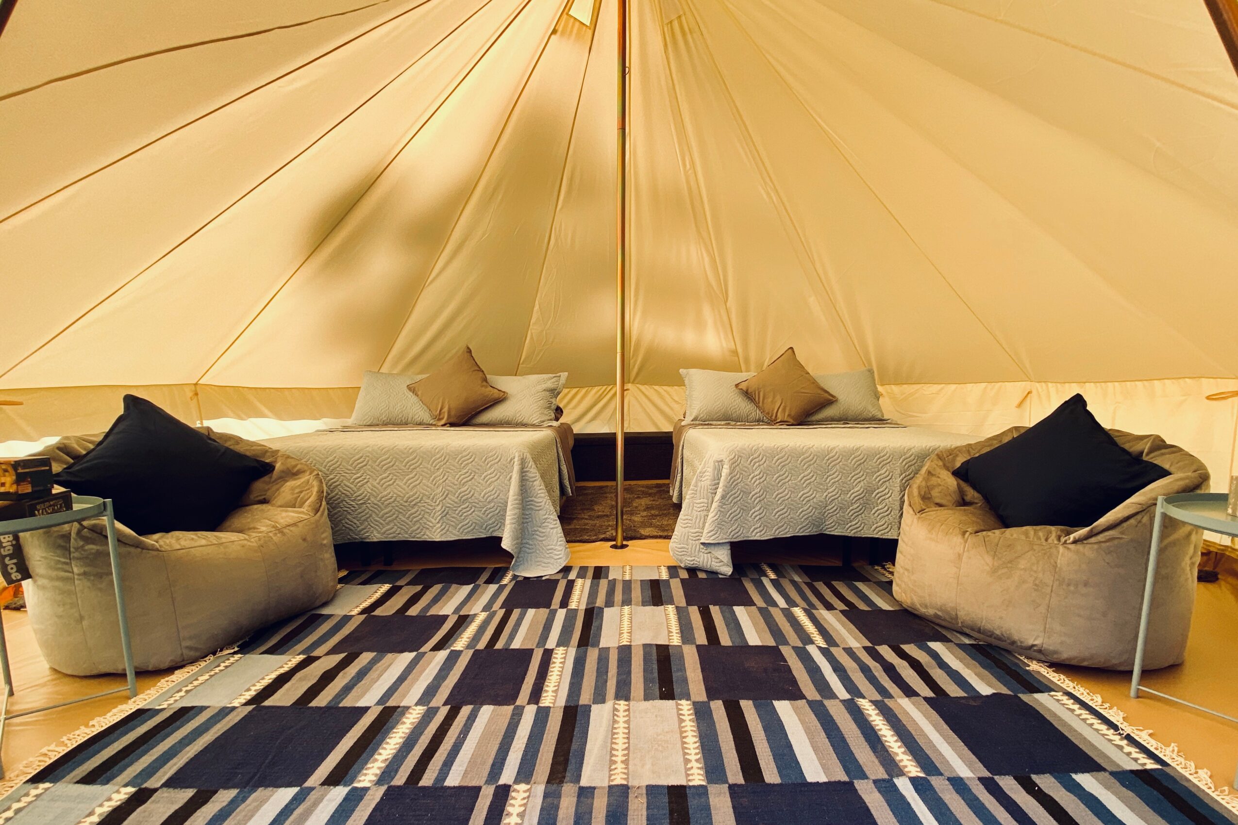 Interior of a tent with two beds, two armchairs, and a patterned rug, with beige and black cushions.