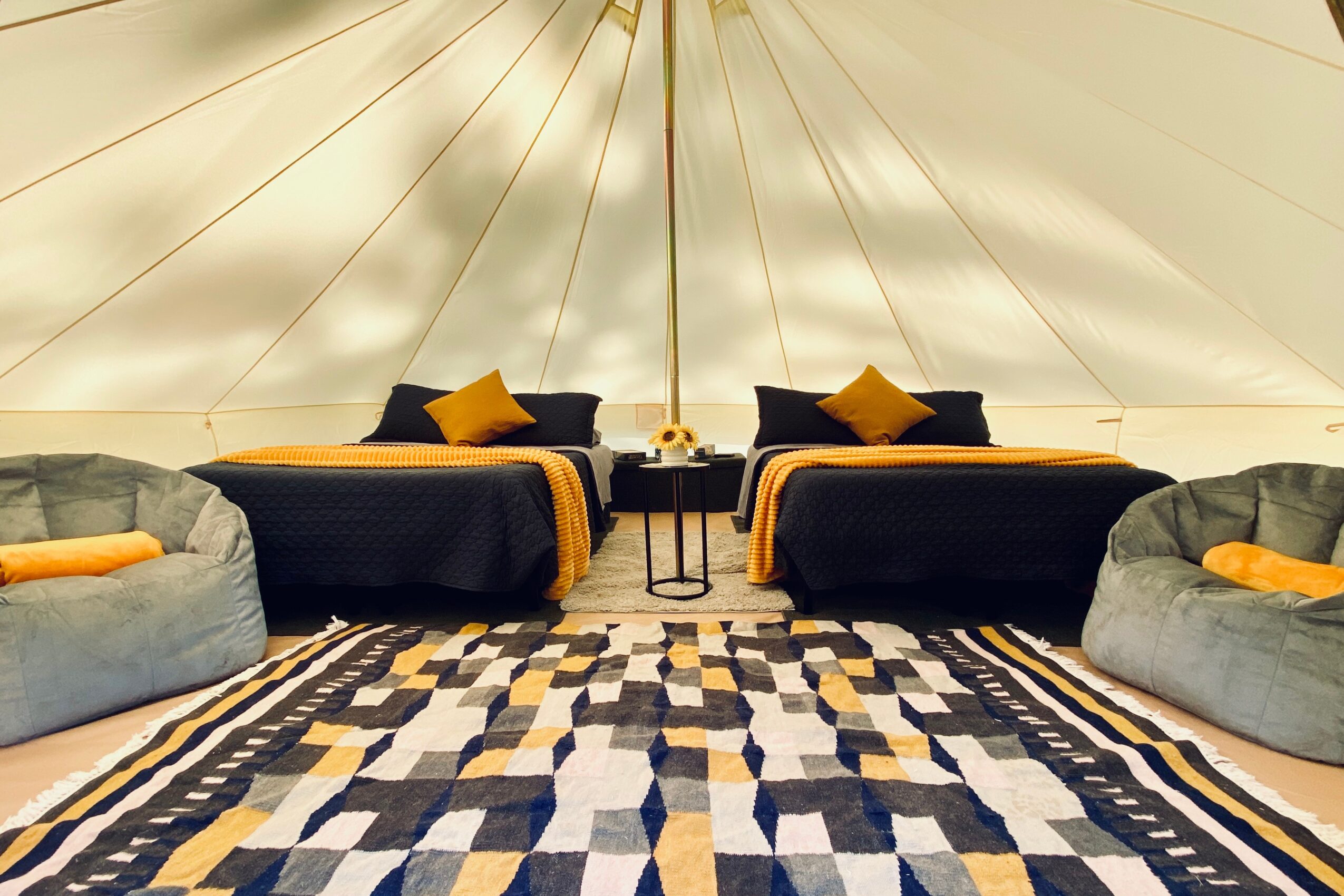 Two beds with black and yellow bedding, two round gray chairs with yellow pillows, and a patterned rug inside a tent.