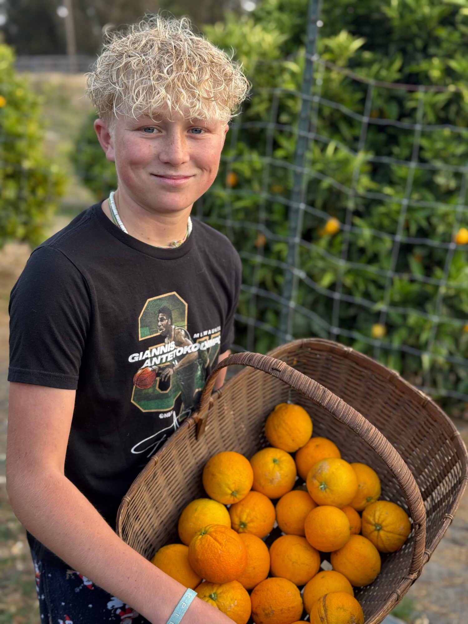 Young person with curly blonde hair holding a basket of oranges outdoors, greenery in background.