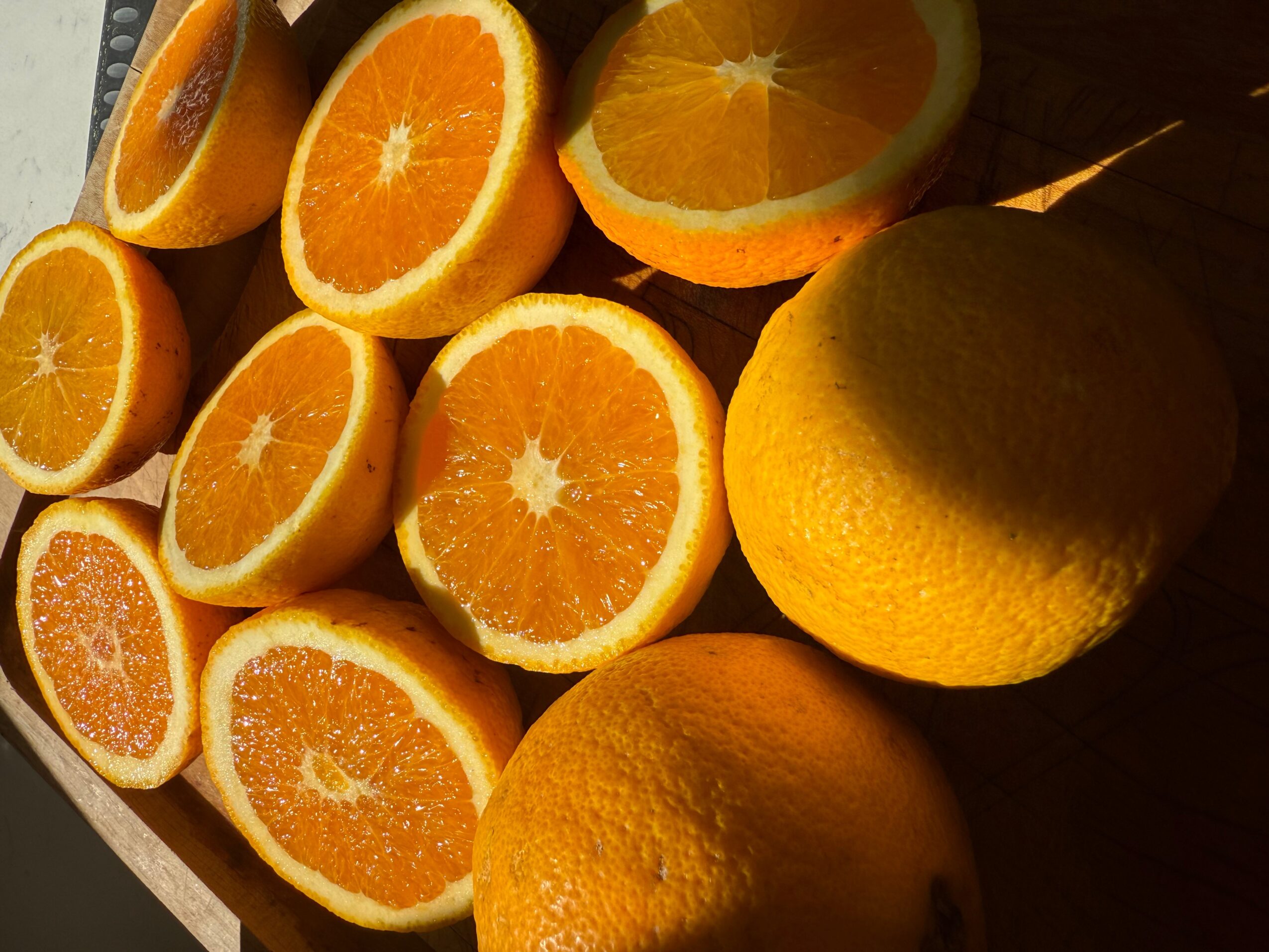 Several halved oranges and two whole lemons on a dark surface, with sunlight casting shadows.