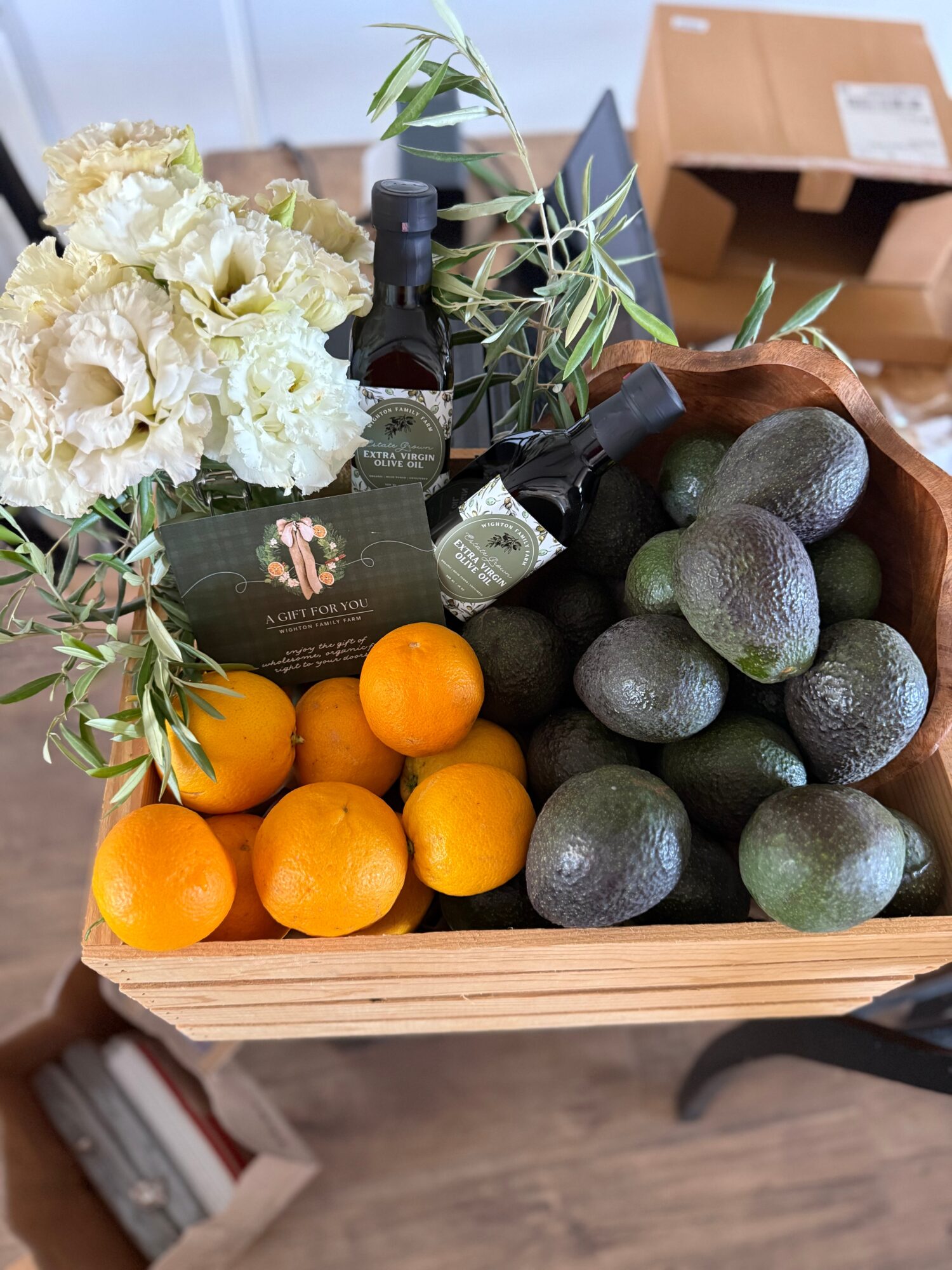 Basket of avocados, oranges, and flowers on a table with boxes in background.
