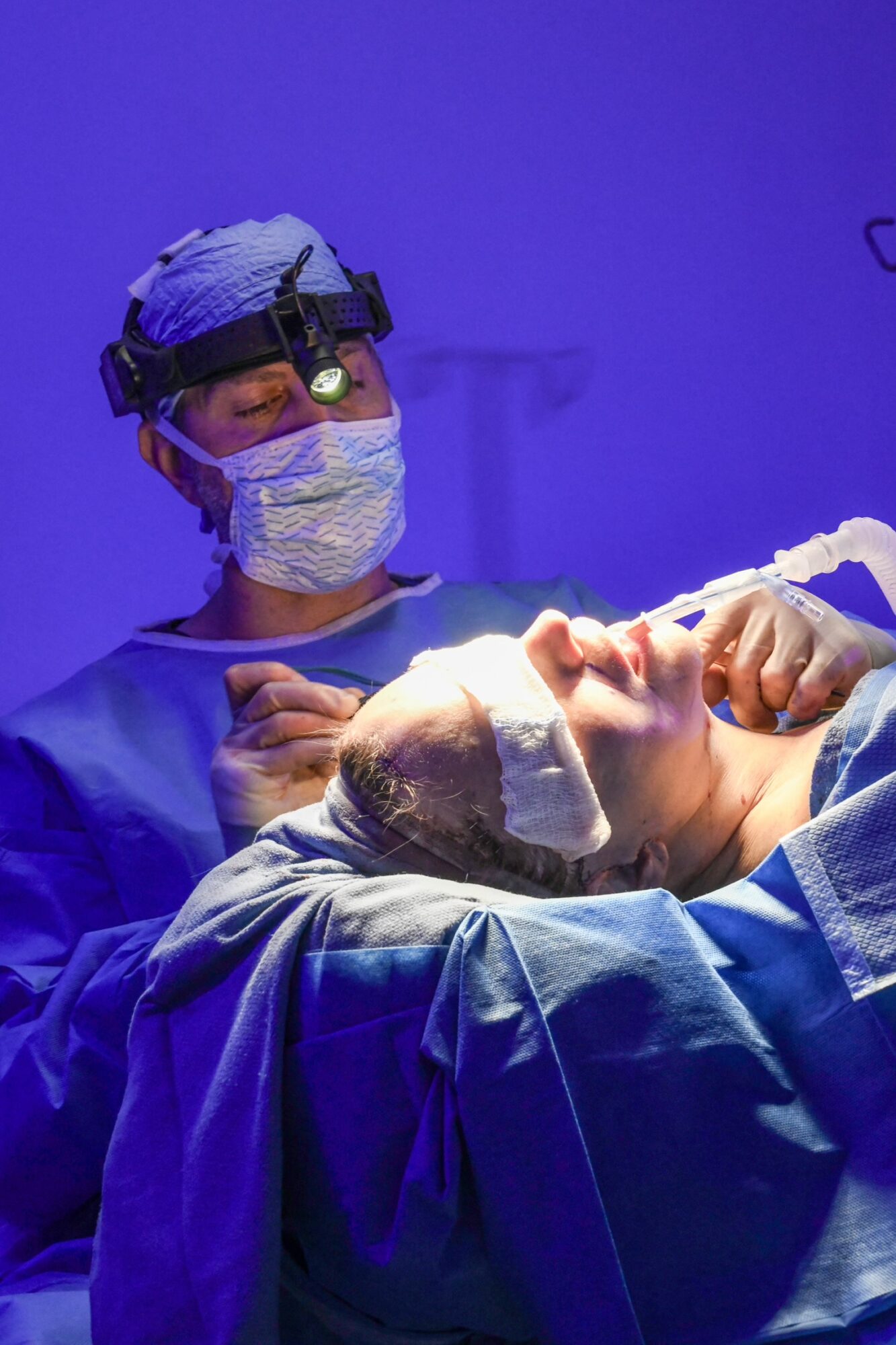 Surgeon wearing a mask, glasses, and headgear performs surgery on a patient in an operating room with blue lighting.