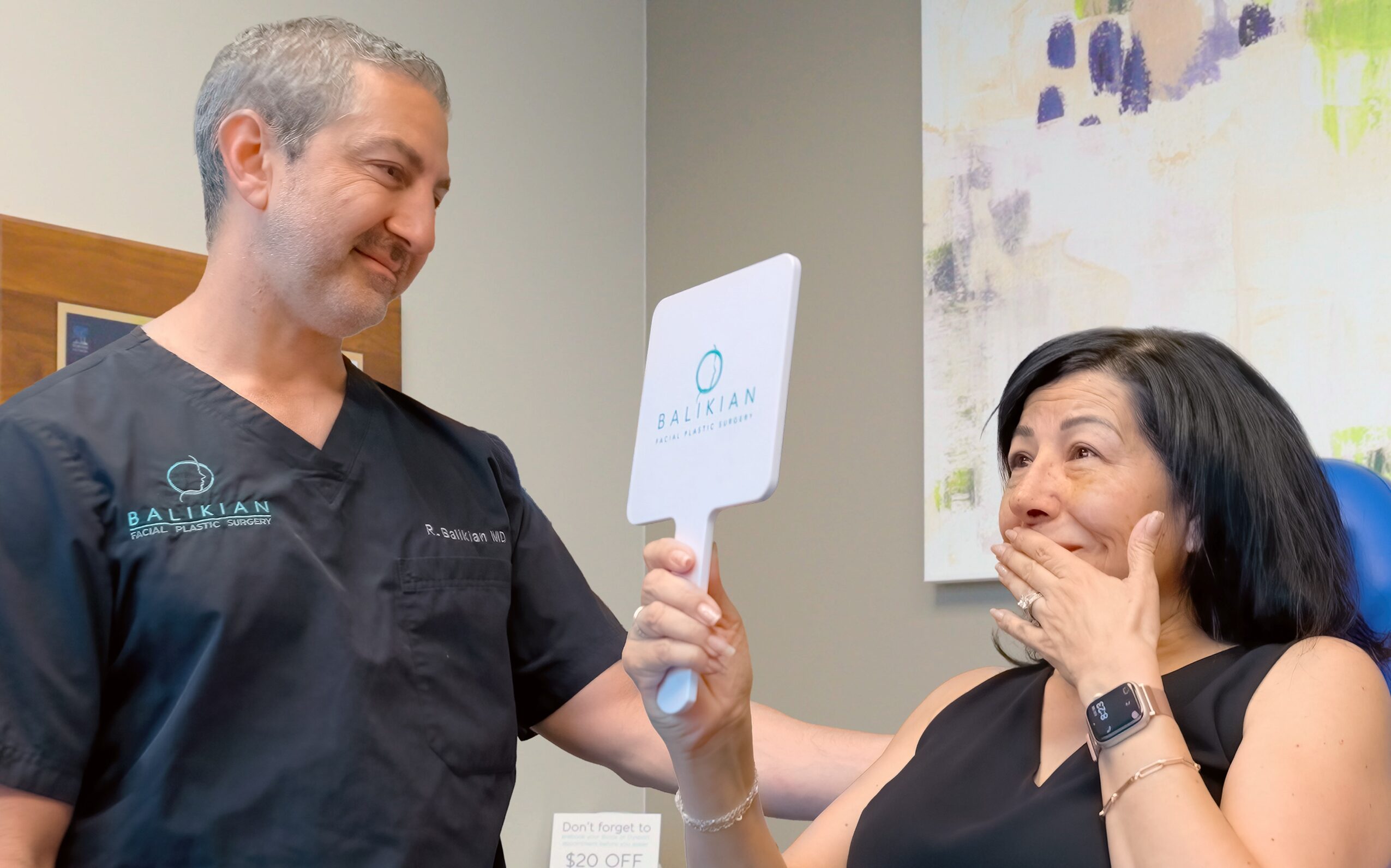 A man in medical scrubs holds a sign while a woman reacts with surprise, covering her mouth with her hand.