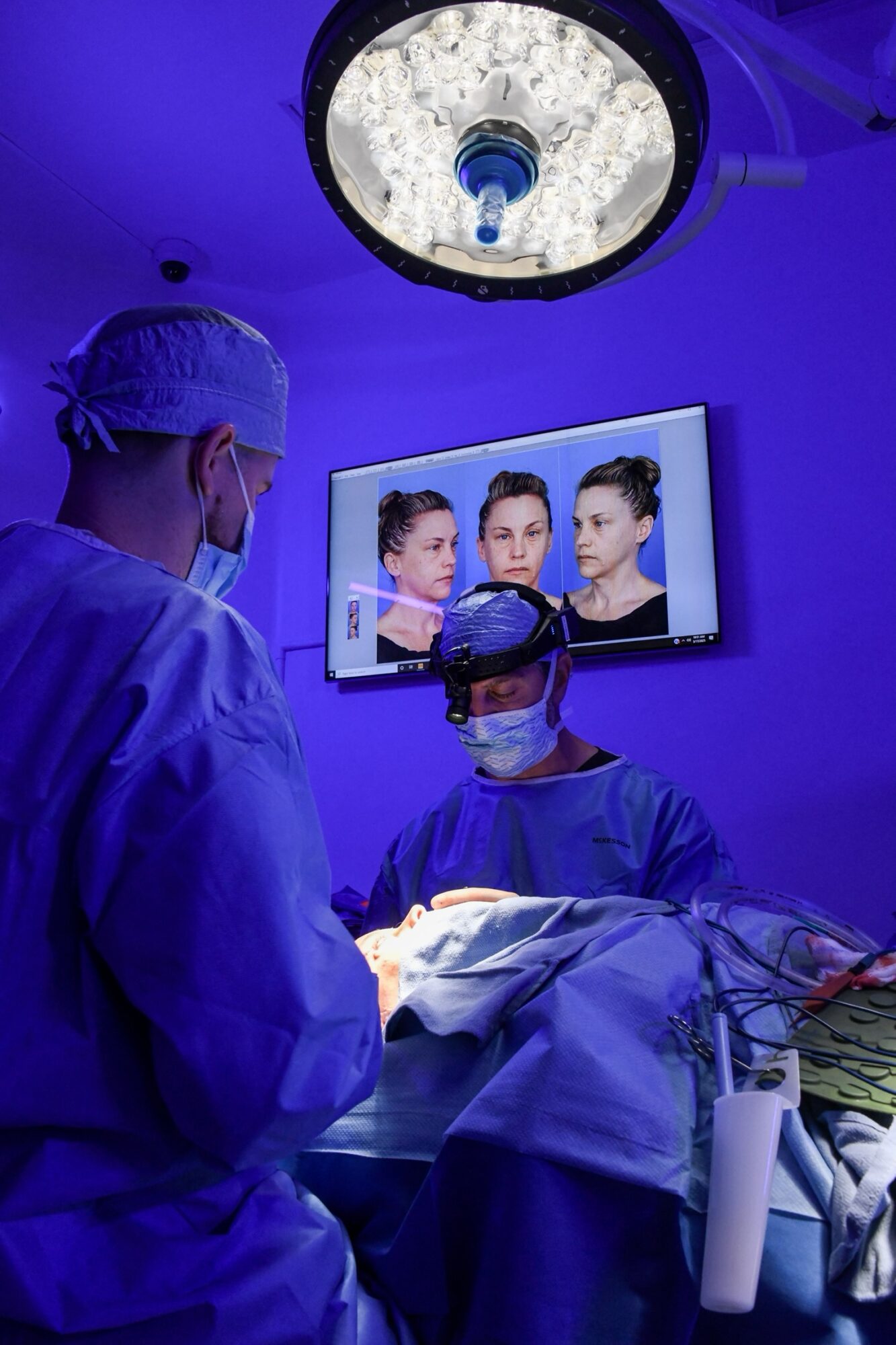 Surgeons in an operating room watch a monitor displaying a woman's face during surgery, with bright surgical lights overhead.