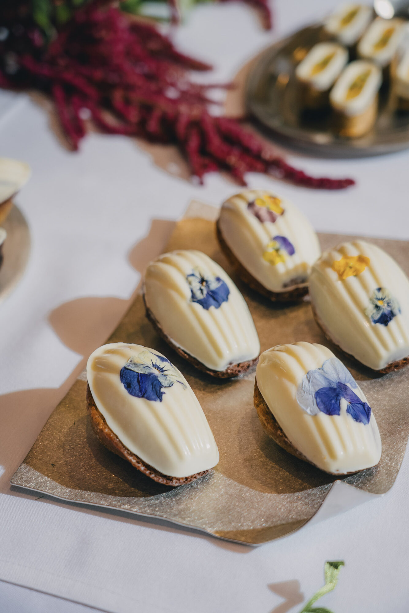 Close-up of decorated cookies with blue, yellow, and purple icing on a brown paper, with a blurred background of more cookies and red flowers.