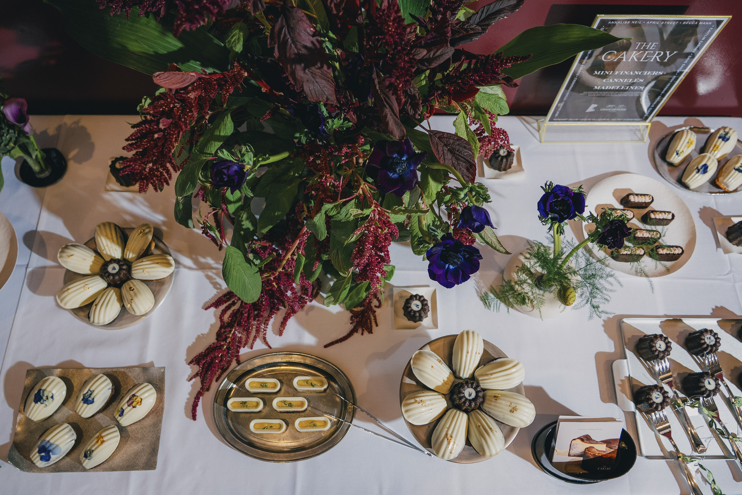 Table with floral arrangements, desserts, and decorative items, viewed from above, with a large central flower arrangement. Alt text word count: 20
