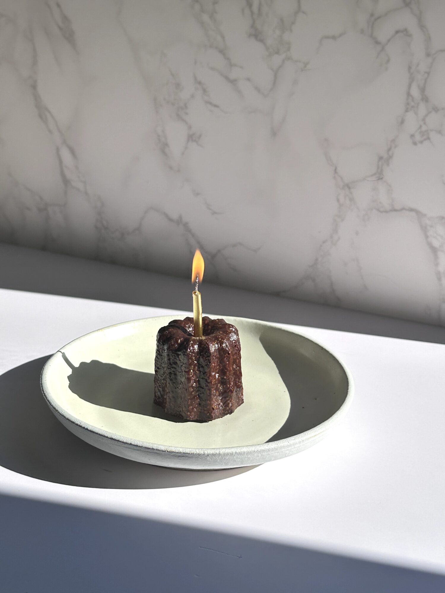 Small chocolate cake with a lit candle on a white plate, shadow cast on surface, marble wall background.