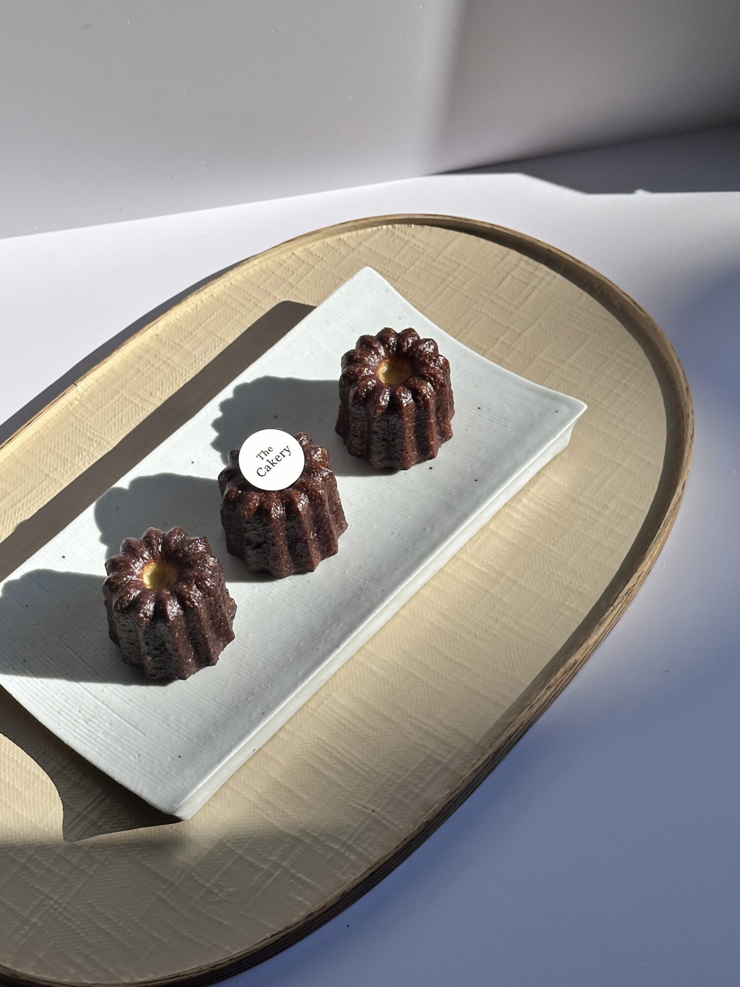 Three chocolate bundt cakes on a white paper towel inside a wooden tray, with shadows cast on a blue surface.