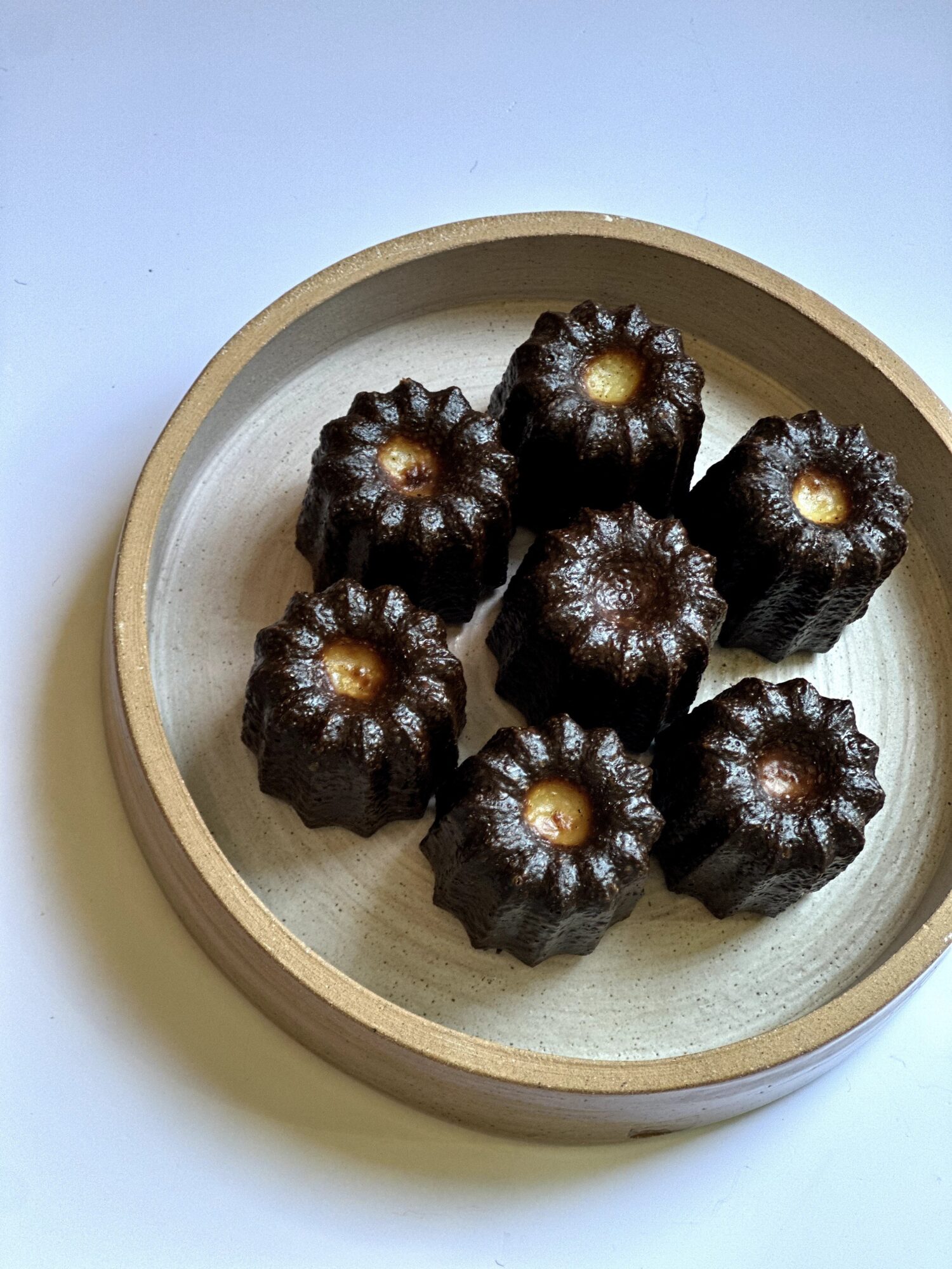 Dark-colored cannelés with golden tops in a round wooden container, viewed from above.
