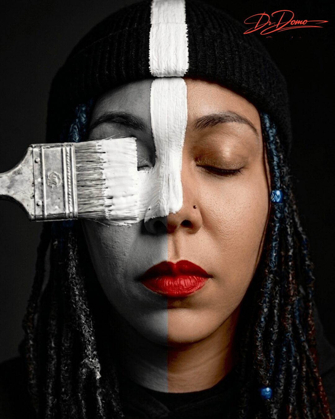 Woman with half face painted white, closed eyes, red lipstick, wearing a black beanie, with a paintbrush on her face.