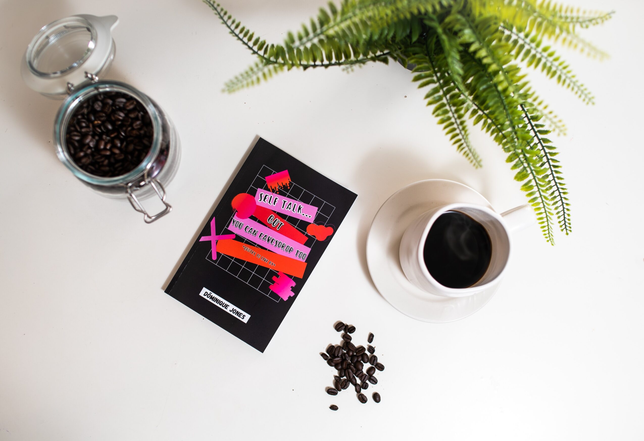 Coffee beans in a jar, a plant, a cup of coffee, and a card on a white surface.