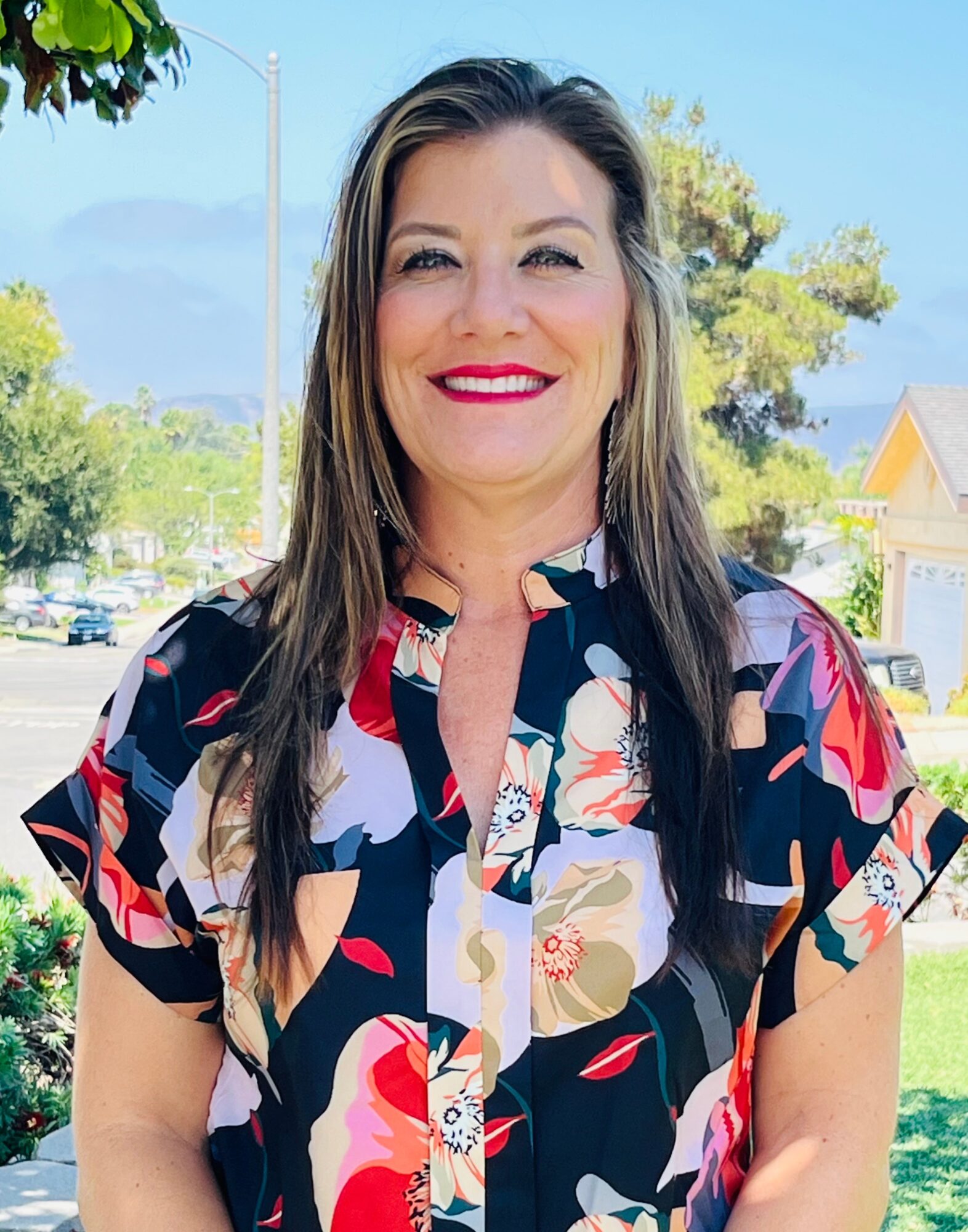 Smiling woman with long dark hair wearing a colorful floral blouse outdoors, trees and buildings in background.