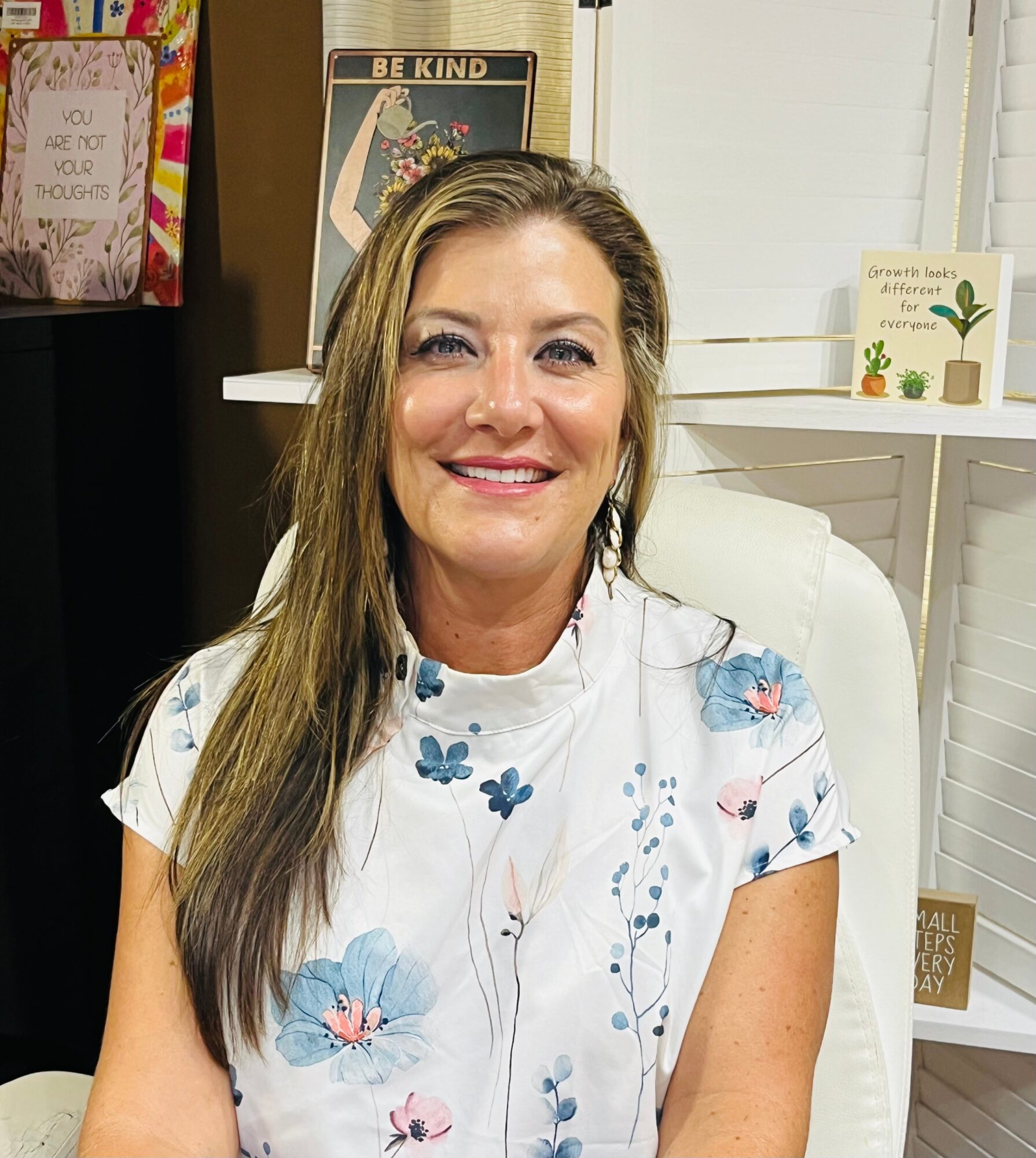Smiling woman with long brown hair wearing a floral blouse, sitting in a room with books and decorations.