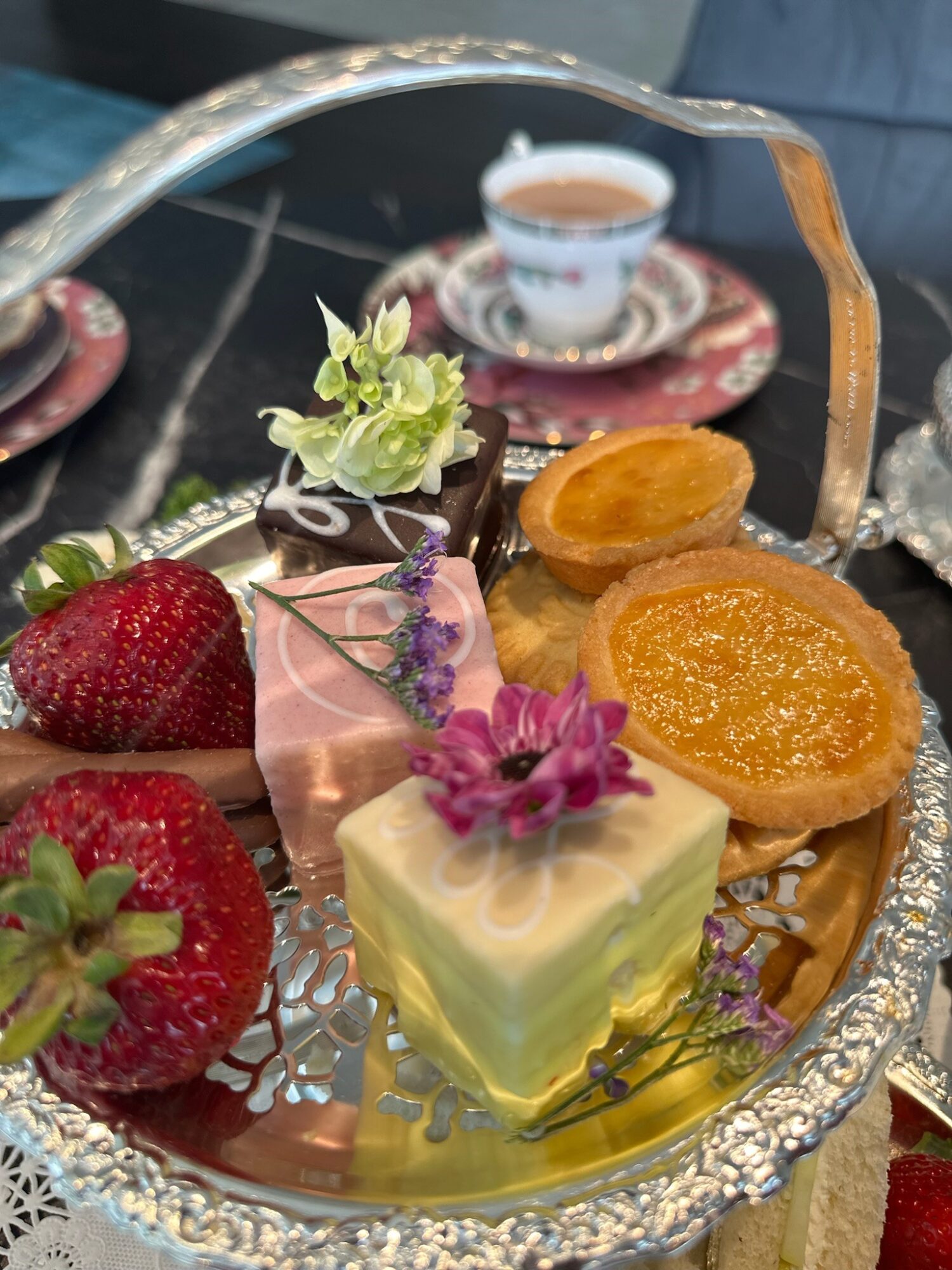 Assorted desserts on a silver tray, including strawberries, a small cake with purple flowers, and fruit tarts, with a teacup in the background.