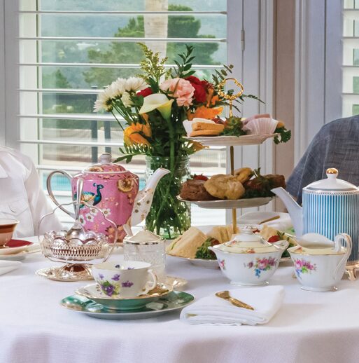 Table with flowers, teapots, cups, and food near window with blinds, sunlight, and greenery outside.