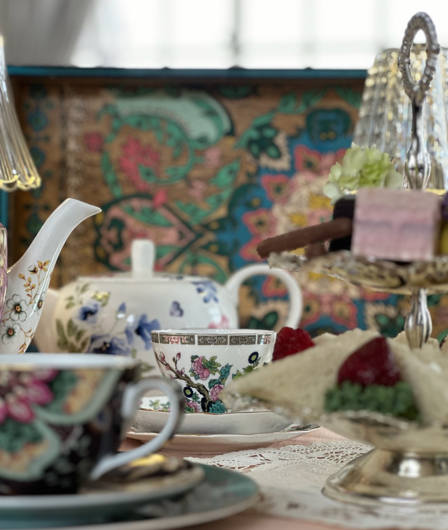 Tea set with cups, teapot, and desserts on a table with a colorful tapestry background.