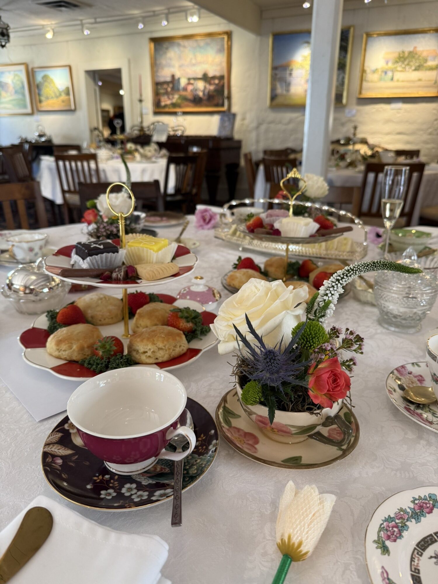 Table set with desserts, flowers, and teacups in a decorated dining room, with paintings on the walls and chairs in the background.