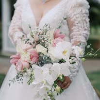 Person in wedding dress holding a bouquet of pink and white flowers, outdoors, with lace sleeves and a deep neckline.