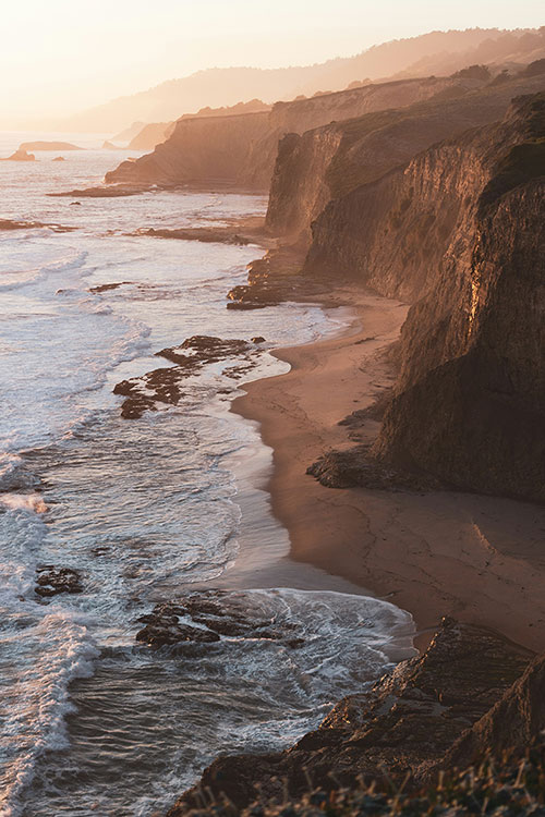 Cliffs and beach along a coastline with waves crashing, under a hazy sky, during sunset or sunrise.