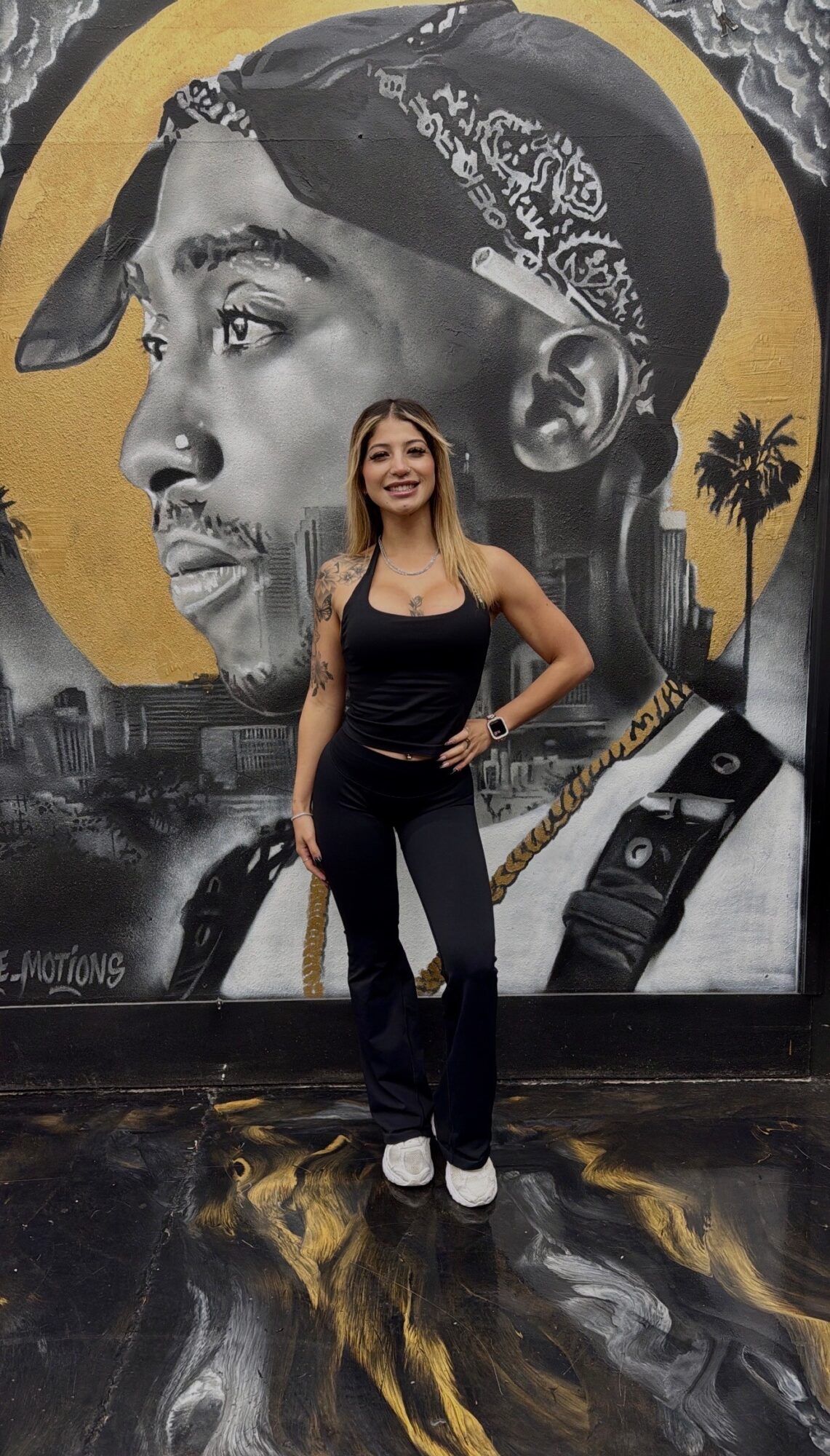 Young woman with long hair, wearing a black tank top and jeans, standing in front of a mural of a man's face with a bandana, palm trees, and cityscape.