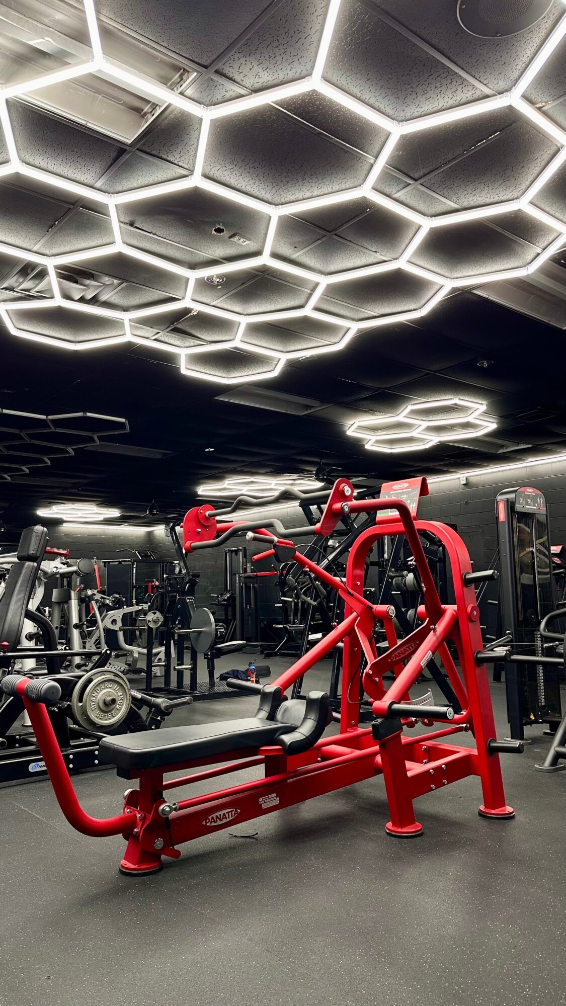 Gym with hexagonal ceiling lights, red workout equipment, and people exercising in the background.