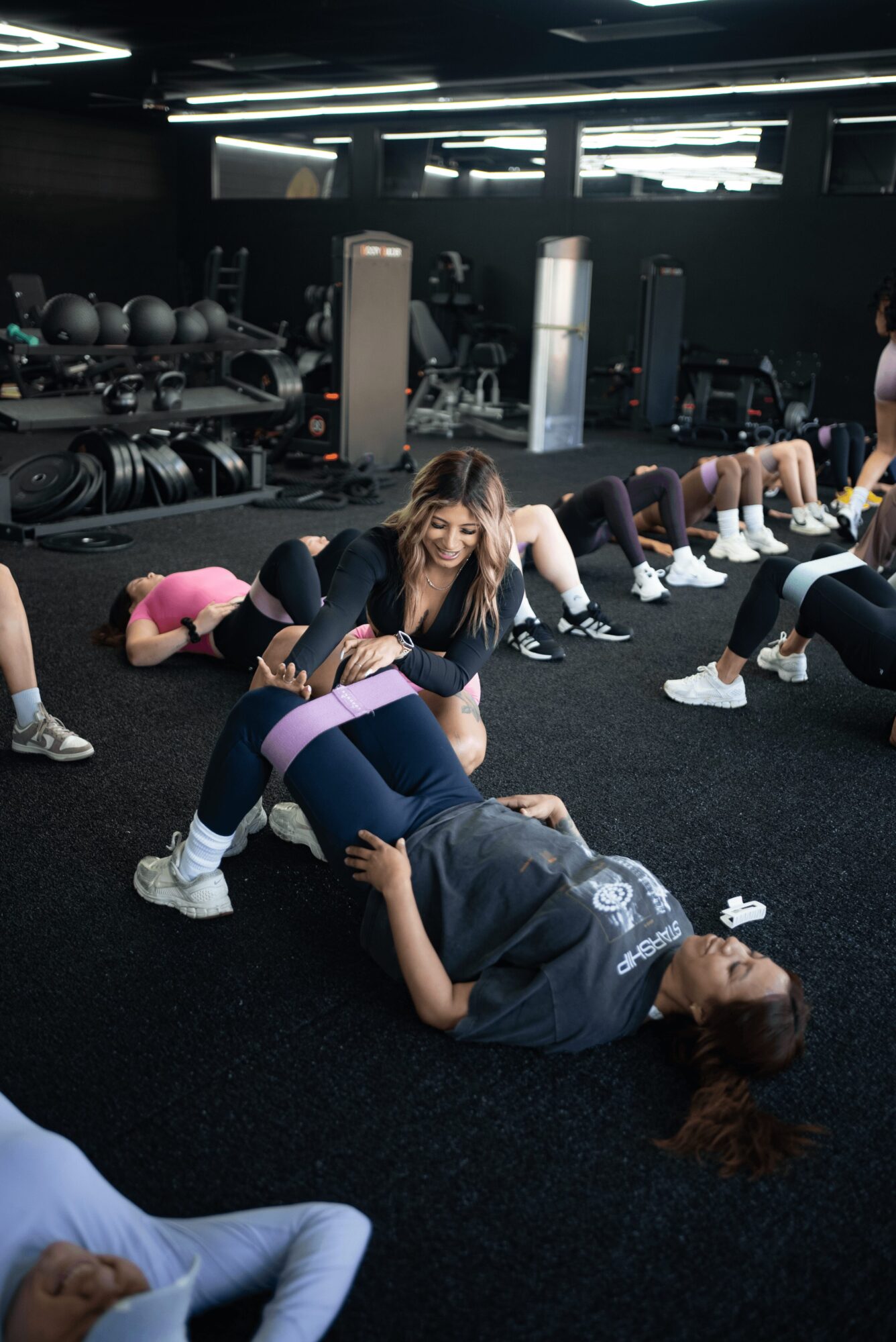 Instructor assisting a person during a fitness class in a gym with exercise equipment and participants stretching or exercising.
