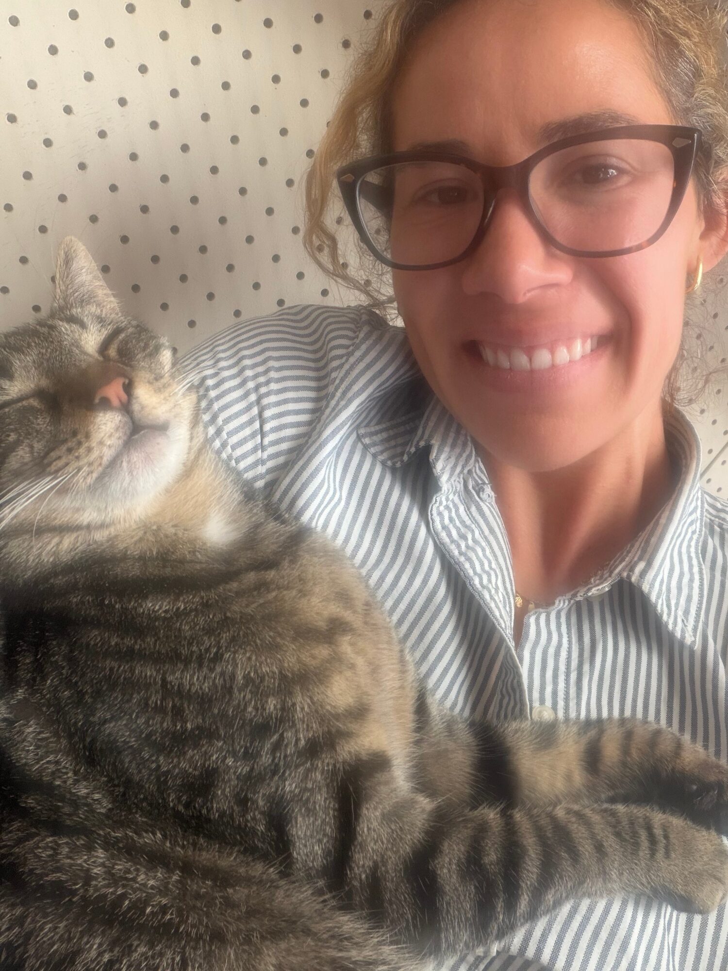 Woman with glasses smiling, lying next to a tabby cat with striped fur, both resting on a polka-dotted surface.