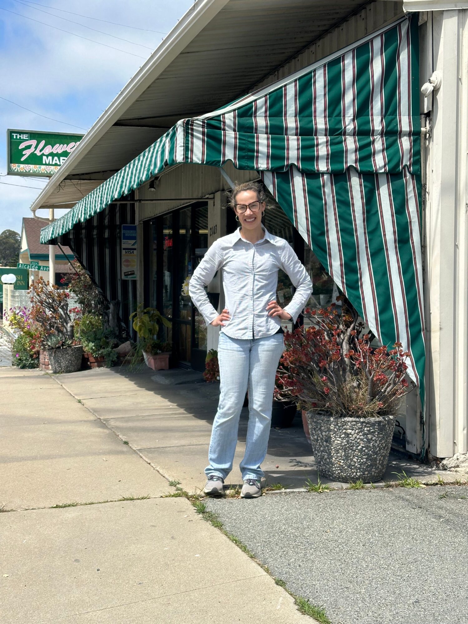 Person standing outside near a building with a striped awning, wearing white clothing, holding a bag, with plants nearby.
