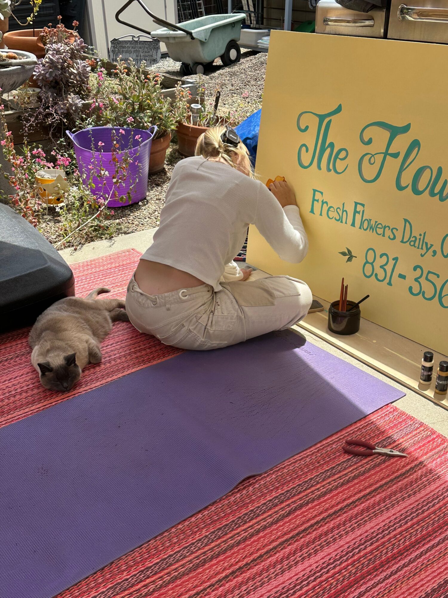 Person sitting on a purple mat, facing away, with a small dog lying nearby on a red and purple mat, in front of a yellow sign.