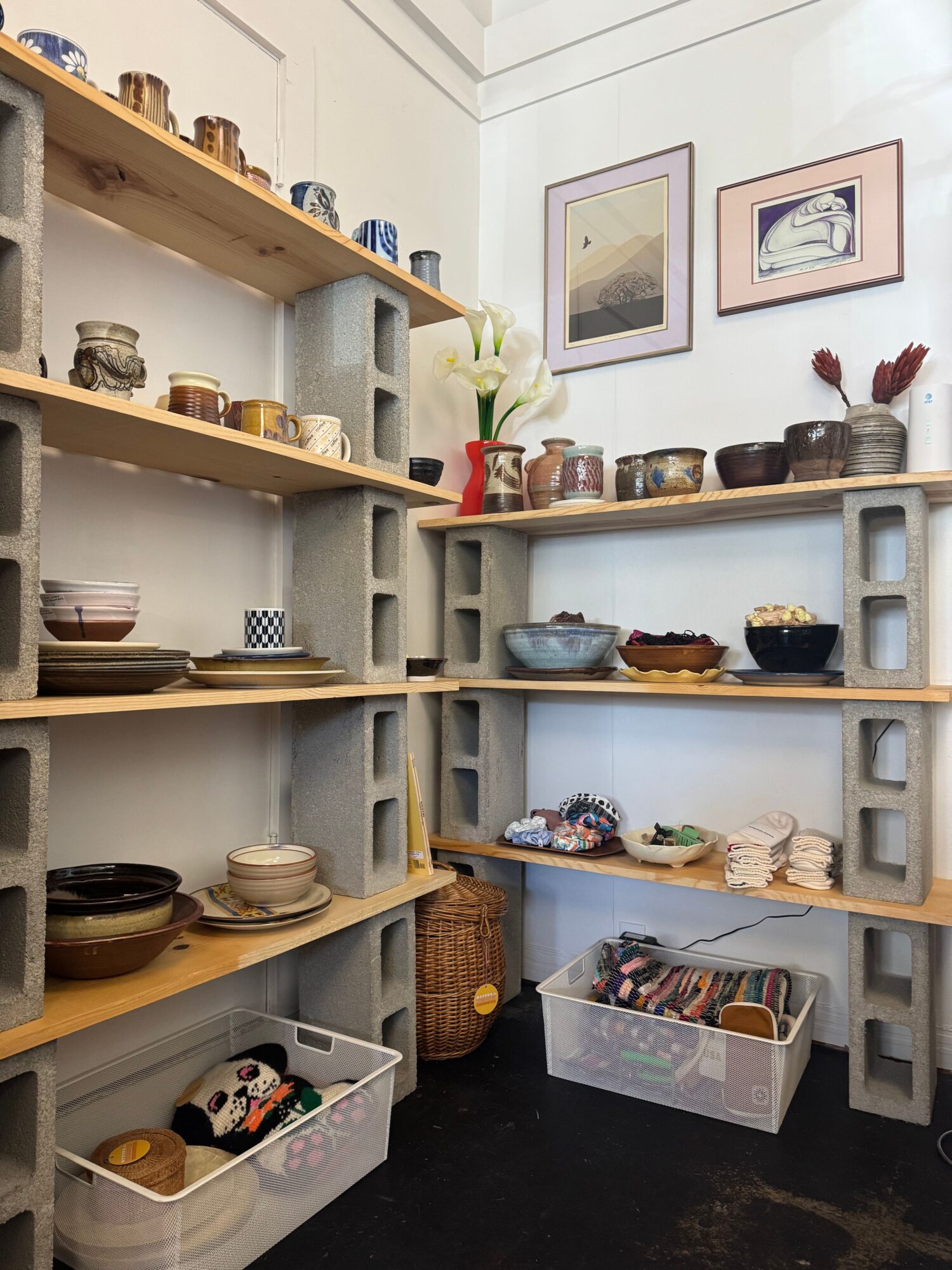 Shelves with jars, bottles, and decorative items, with framed pictures on the wall behind, and a clear plastic container on the right.