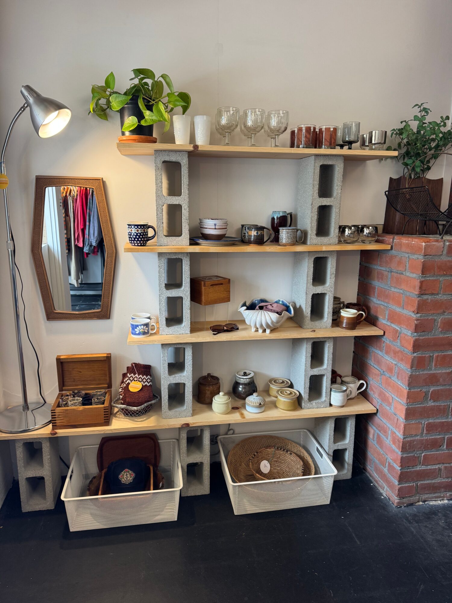 White shelving unit with kitchen items, plants, and a mirror, against a brick wall and dark floor.