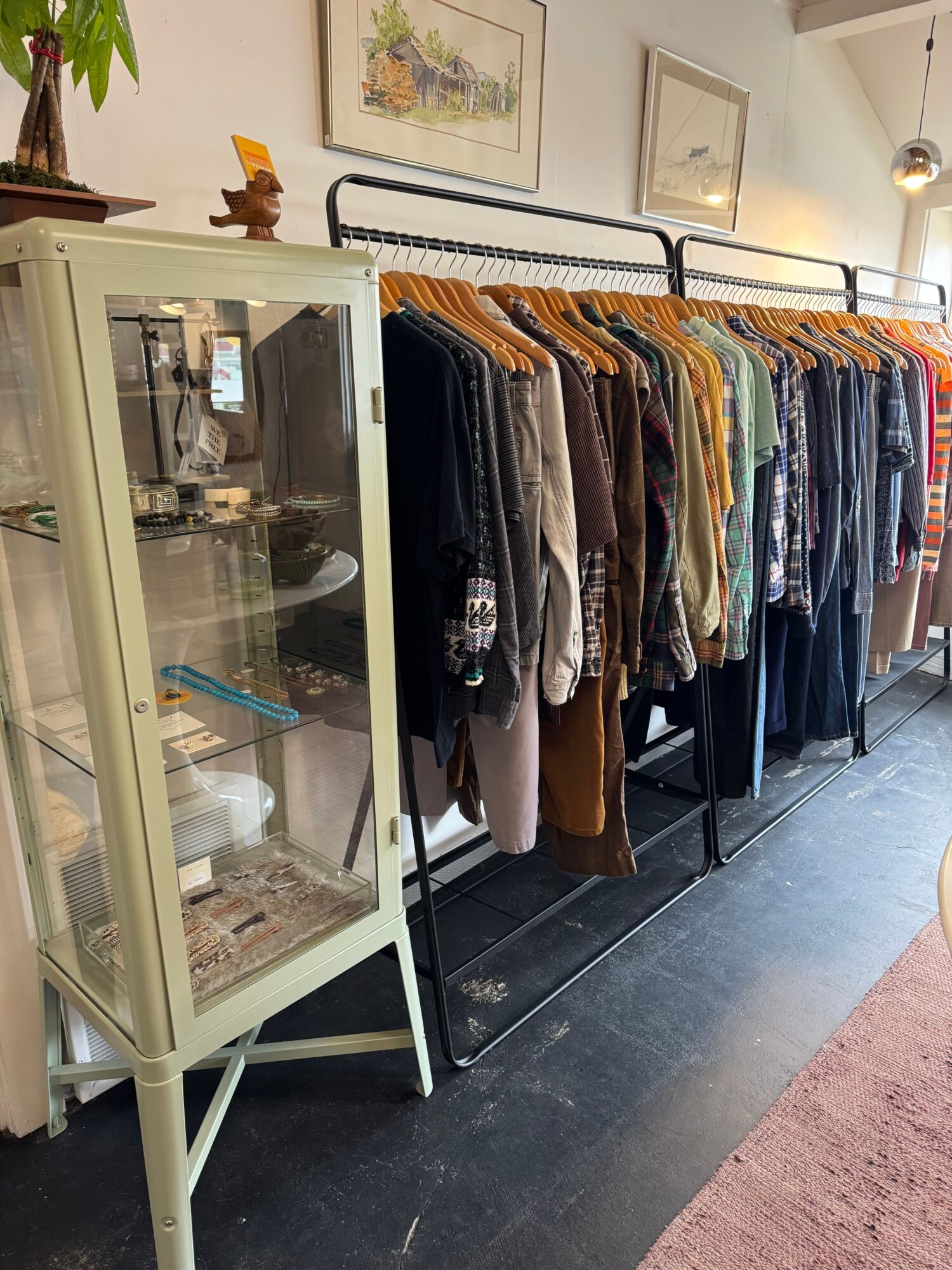 Clothing rack with various garments, glass display case, and framed artwork on wall in a store or gallery.