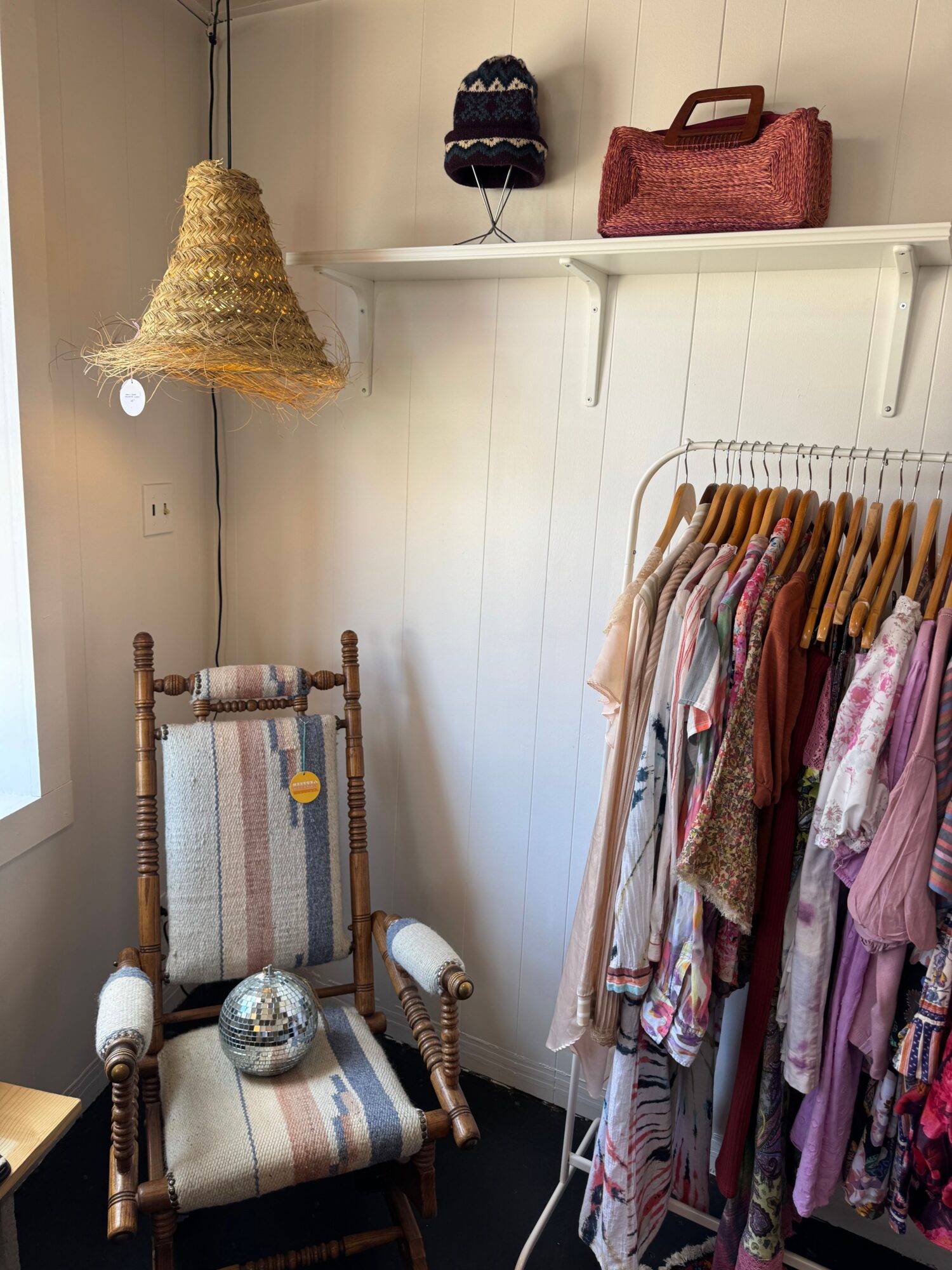 Clothing rack with pink and patterned fabrics, a chair with a white cushion, and a hanging woven decoration in a room.