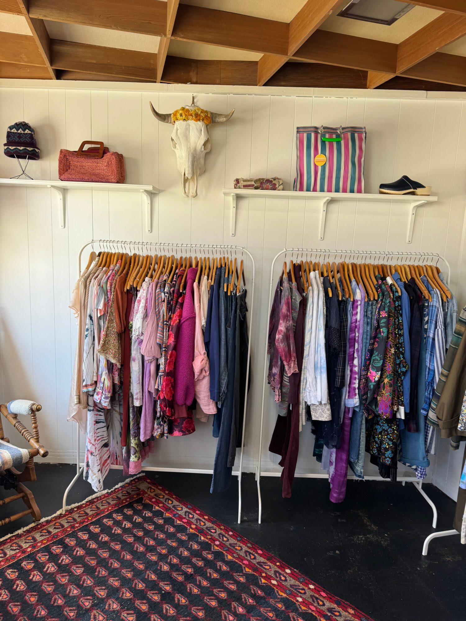 Clothing racks with colorful garments in a store, with a patterned rug on the floor and shelves on the wall.