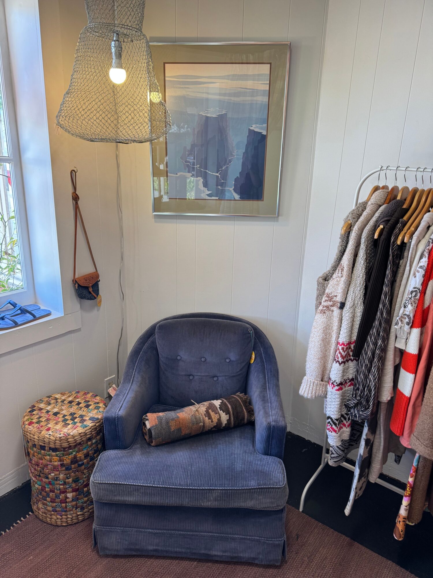 Upright blue armchair with a pillow, next to a woven ottoman, in a room with a framed picture and clothing rack.