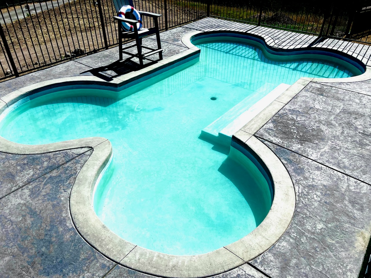Empty swimming pool with a person sitting on a bench nearby, surrounded by a fence and concrete surface.
