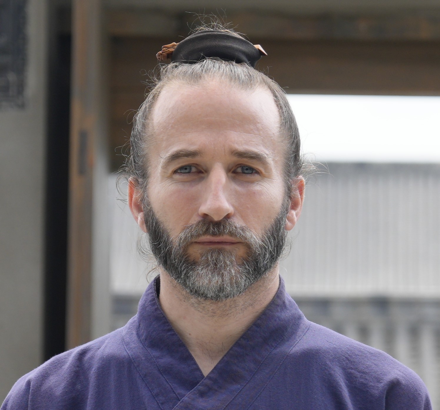 Man with a beard and hair tied back, wearing a dark purple traditional garment, standing outdoors with a building in the background.