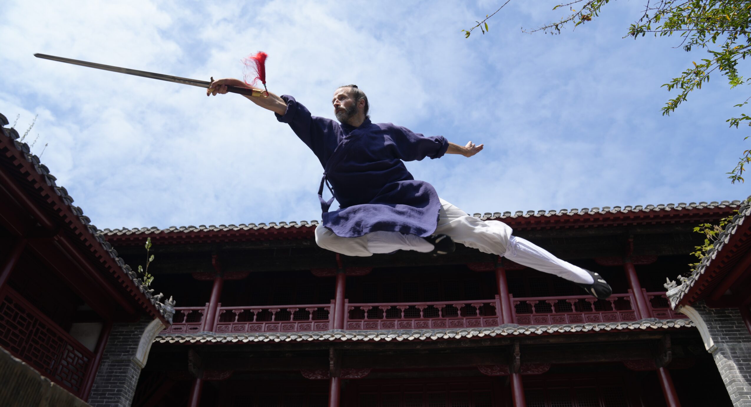 Person performing a martial arts move mid-air in a traditional courtyard with buildings and a blue sky.