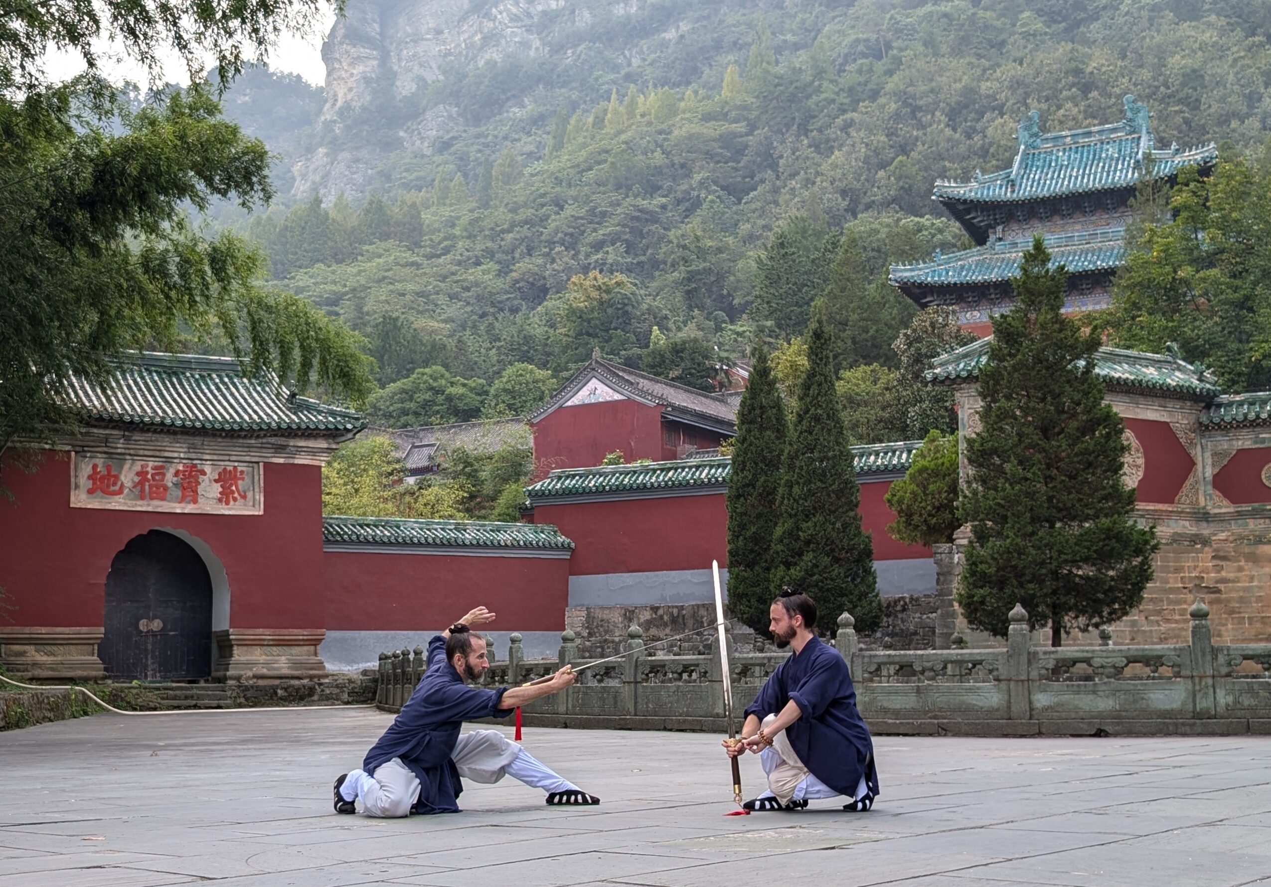 Two people in traditional clothing are playing a game with sticks outdoors, with traditional buildings and trees in the background.