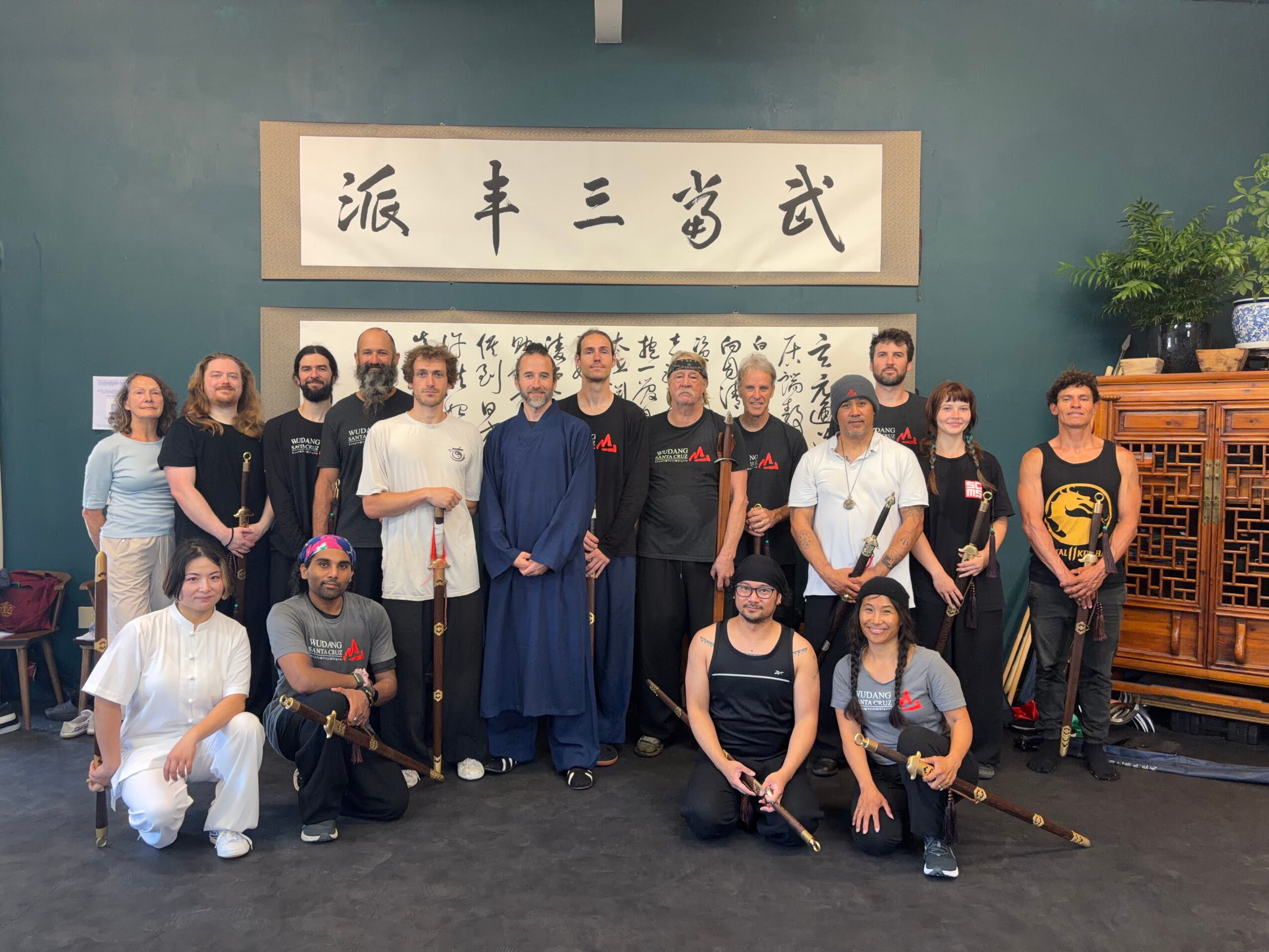 Group of people posing indoors with Chinese calligraphy and decorations, some holding swords, in front of a blue wall.
