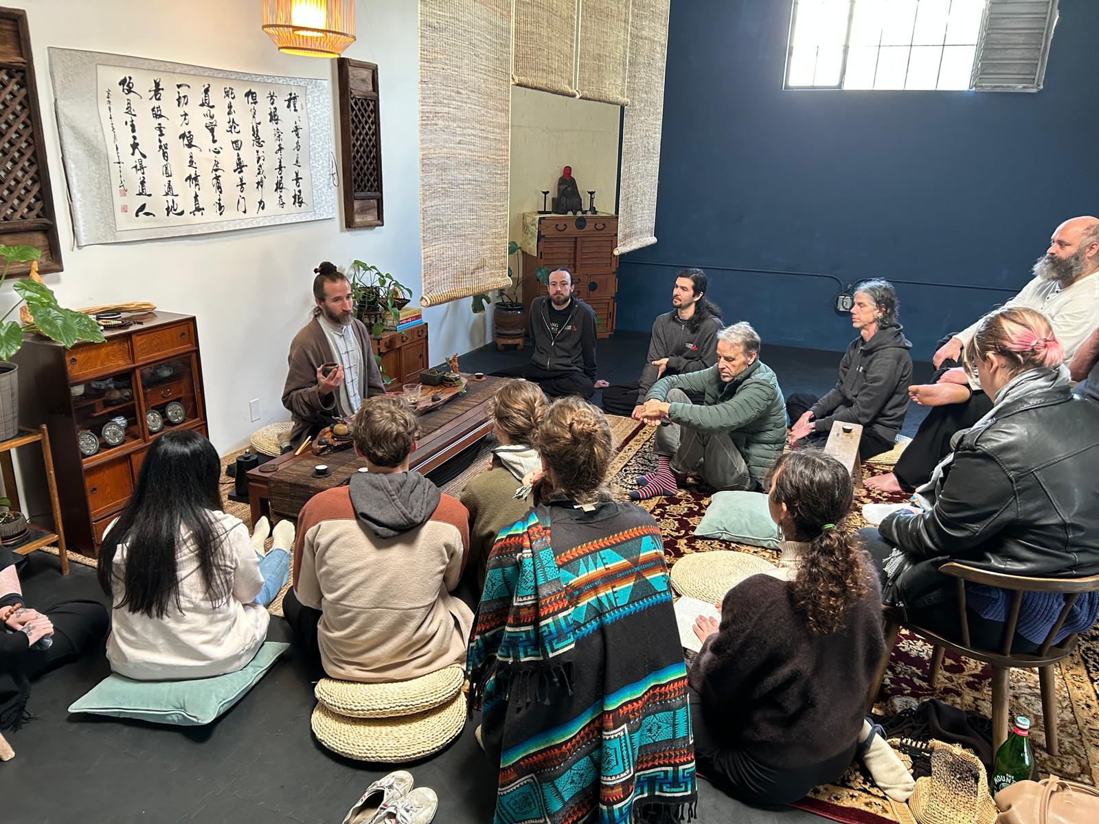 Group of people sitting on cushions and chairs in a room with traditional Asian decor, listening to a speaker.