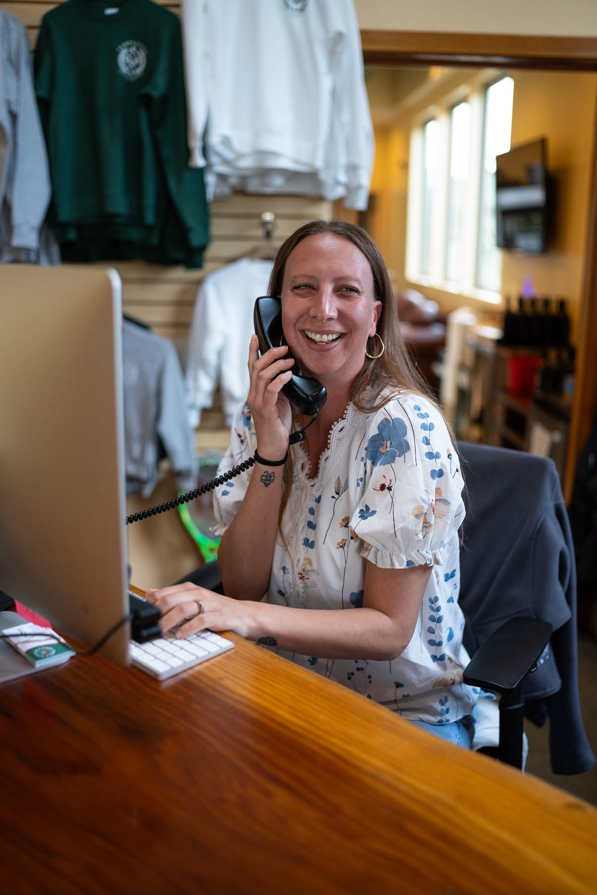 Woman smiling and talking on phone at desk in clothing store, with clothes hanging behind her.