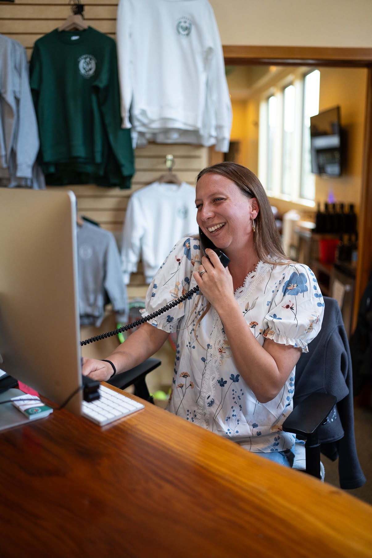 Woman smiling and talking on the phone at a desk with a computer, in a room with sports jerseys hanging on the wall.