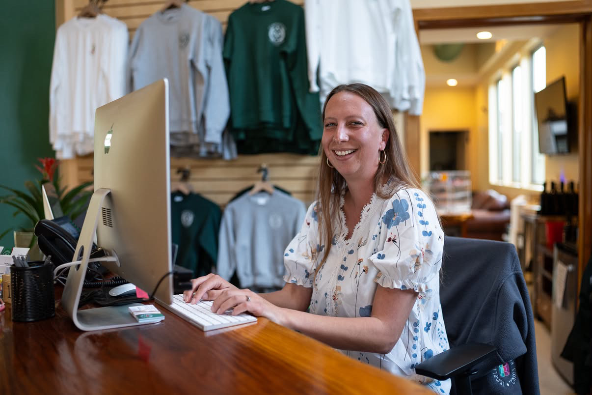 Woman smiling at computer in clothing store with shirts hanging behind her.