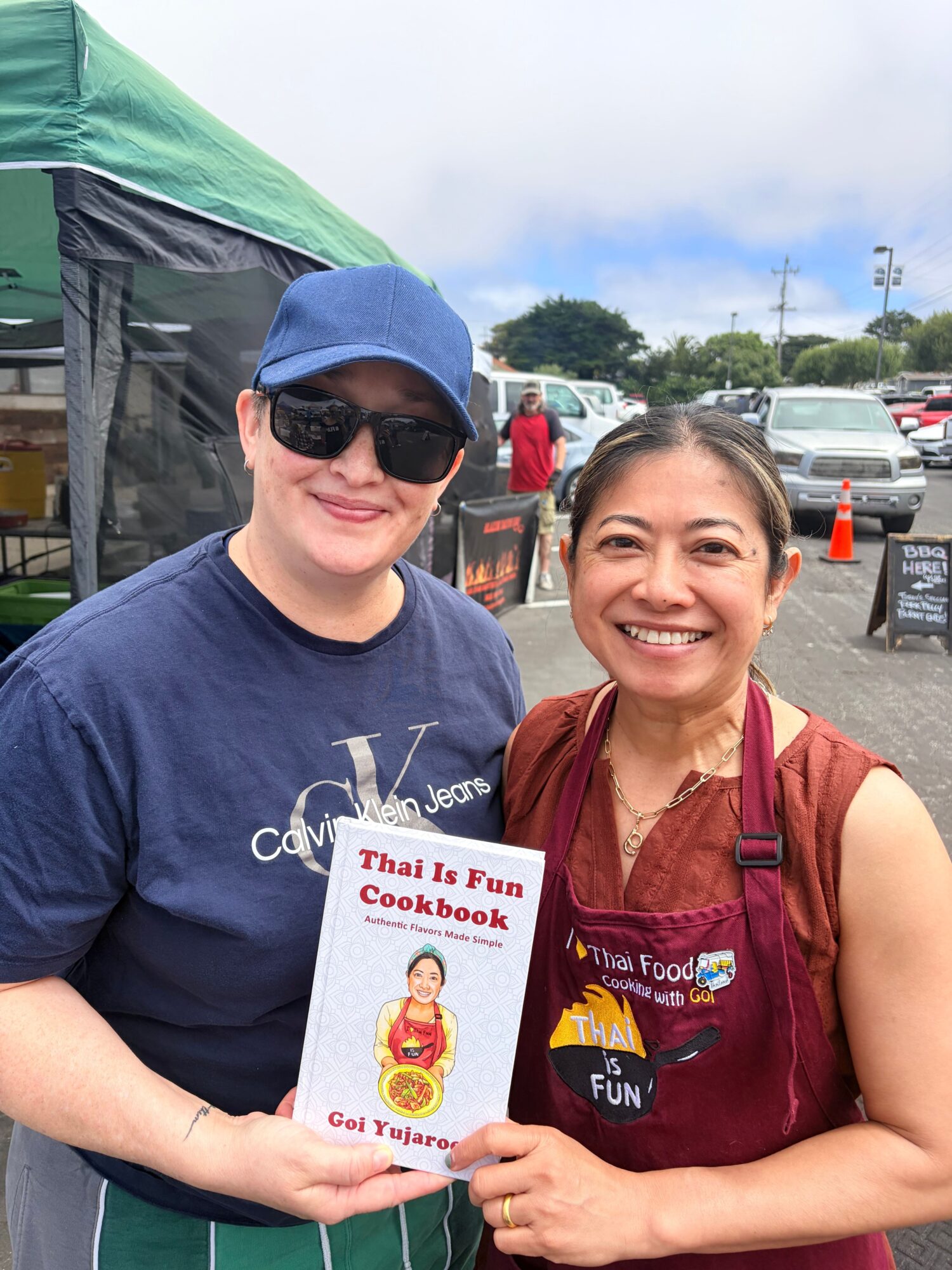 Two women smiling outdoors, one holding a colorful book, with cars and a green tent in the background.