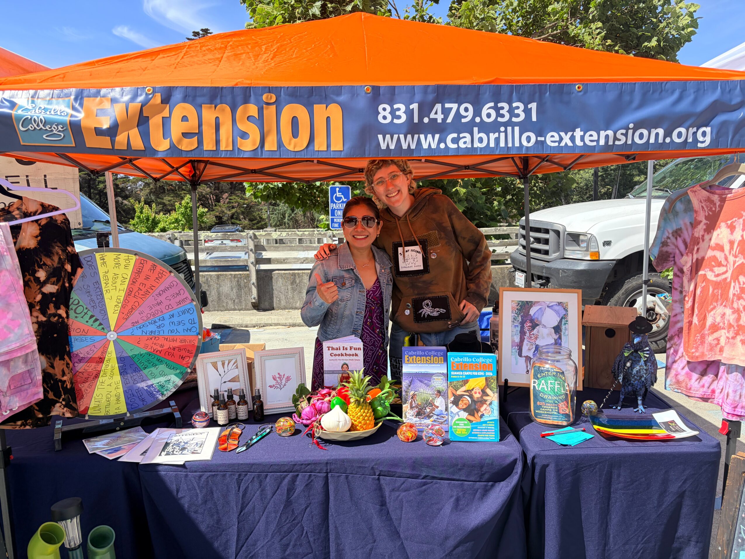 Two women stand behind a table under an orange canopy with 'Extension' and contact info, outdoors with trees and cars.